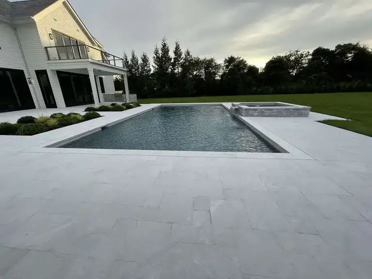 Rectangular pool and raised stone spa on a pale stone terrace beside a white shingle house, lawn and evergreens beyond