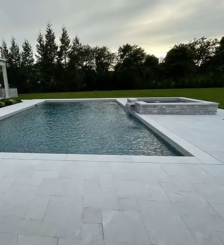 Pool and raised stone spa edged in pale coping, a wide bluestone patio in the foreground and evergreens at dusk