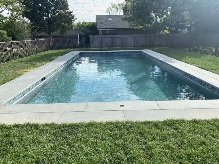 Pool with stone coping and a mosaic tile band at the waterline, set into a green lawn inside a black fence