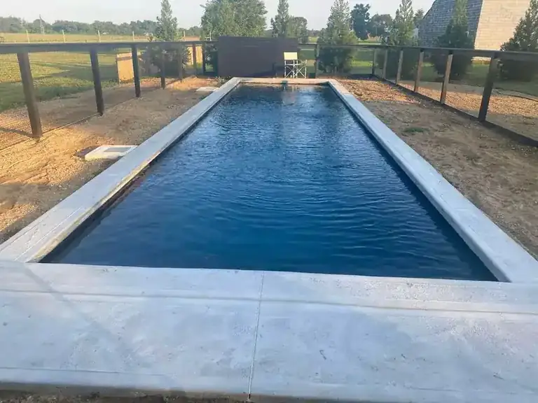 Long, narrow lap-style pool in dark water with wide pale coping, fencing and open fields beyond