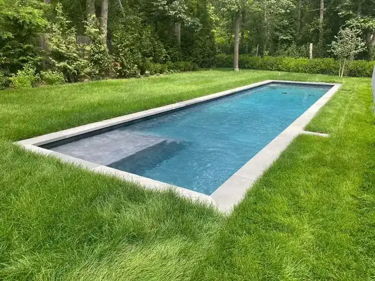 Long rectangular pool with pale stone coping set into a lawn, woods behind