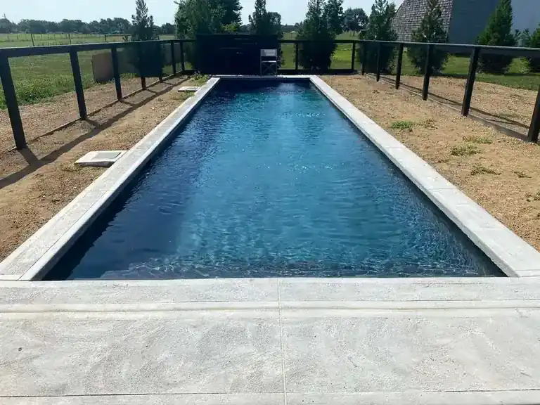 Long pool in dark water with pale stone coping and a poured concrete deck, a black fence and open fields behind