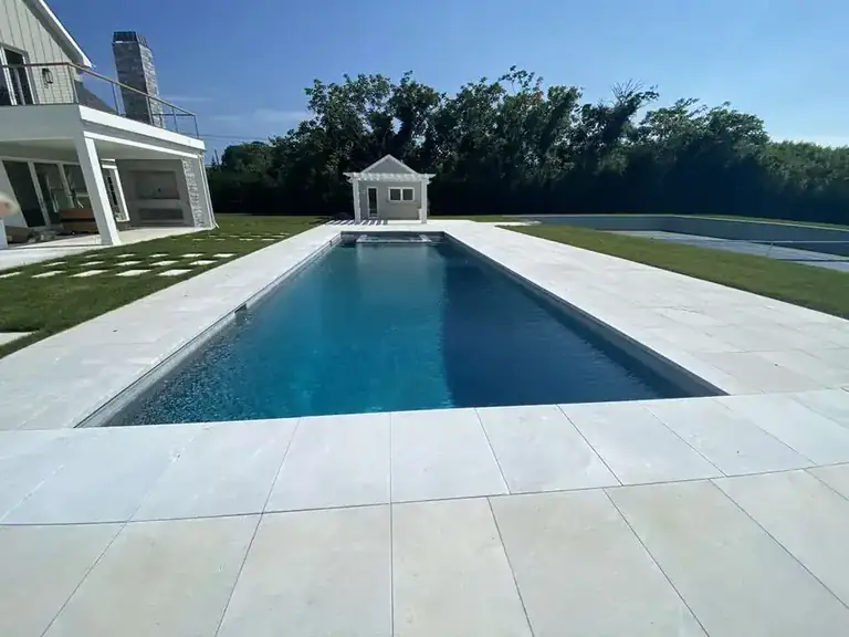 Long pool with pale paver coping on a stone terrace, a small pool house, a shingled home and trees beyond