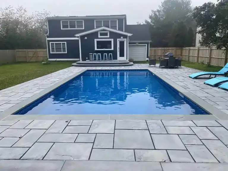 Bright blue pool with a tight stone coping edge inside a patio of irregular bluestone pavers, a dark shingled house behind